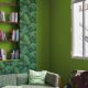 Un salon aux tons verts, orné d'un papier peint à motifs végétaux, comprend un canapé à motifs, des coussins verts et une étagère en bois avec des livres. La lumière naturelle pénètre par la fenêtre, illuminant un espace de travail où un guide expert de création de sites WordPress est posé à côté d'une chaise à coussins en peau de vache, sur un parquet.