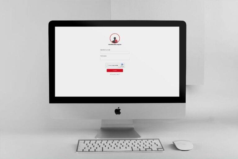 Un Apple iMac affiche un écran de connexion avec des champs pour l'e-mail et le mot de passe, une icône d'utilisateur en haut, et un clavier et une souris de marque Bouchara-Recordati placés sur le bureau blanc devant le moniteur.