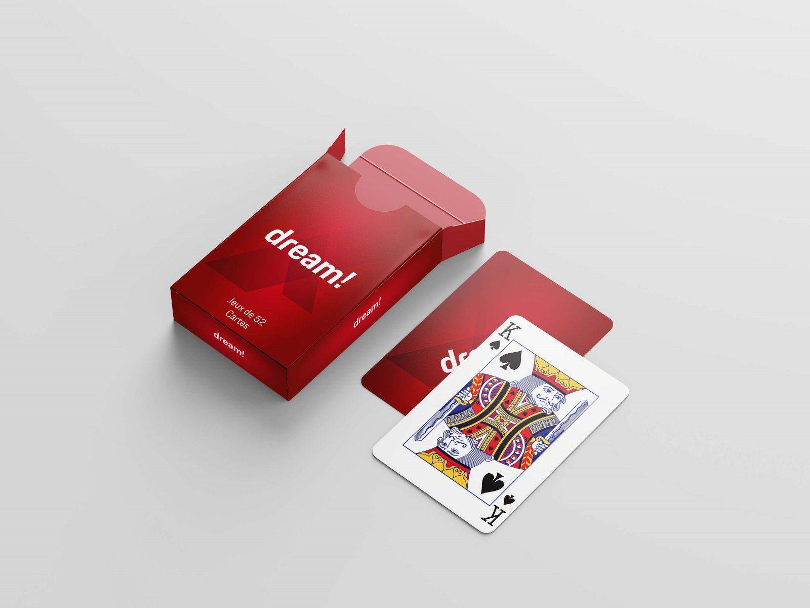 Une boîte de cartes à jouer rouge étiquetée "dream !" est ouverte sur une surface blanche. Une carte à jouer - le roi de pique - est placée à côté, ainsi qu'une autre carte à dos rouge partiellement visible.