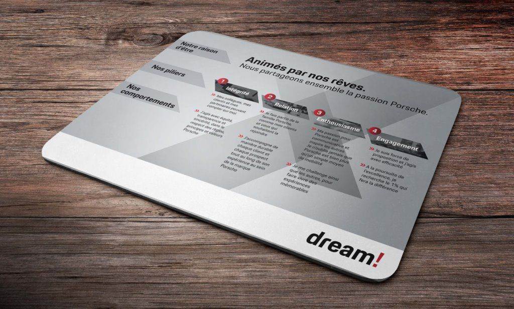 Sur une table en bois, un tapis de souris affiche un diagramme en français représentant les cinq piliers de Porsche : Intégrité, Passion, Énovation, Excellence et Engagement, et le mot dream ! dans le coin inférieur droit.