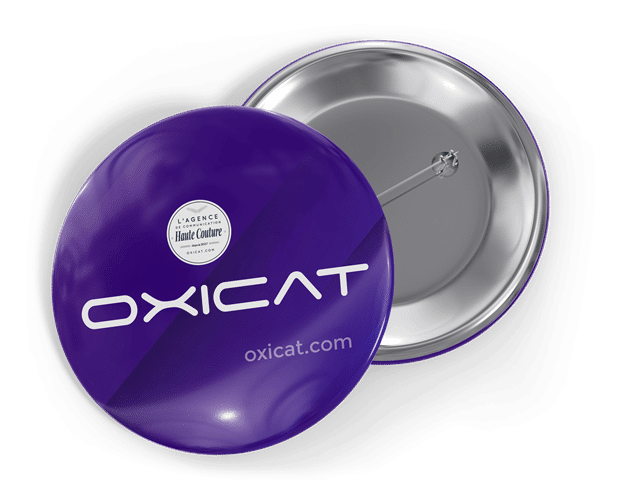 Un badge rond, de couleur violette, sur lequel sont inscrits OXICAT et oxicat.com en texte blanc. Le badge est présenté face vers le haut et face vers le bas, révélant une épingle en métal au dos.