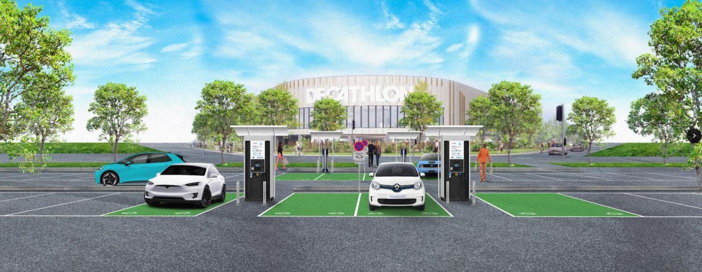 Un parking moderne avec des bornes de recharge pour véhicules électriques, deux voitures en train de charger et un magasin Decathlon en arrière-plan. Des personnes se dirigent vers l'entrée et des espaces verts avec des arbres entourent le parking.