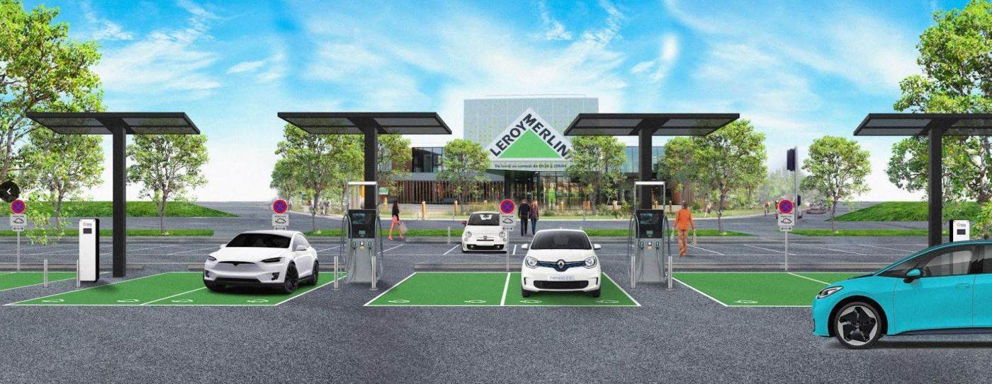 Une station moderne de recharge de véhicules électriques avec quatre voitures garées et rechargeant dans des emplacements marqués en vert, située devant un magasin Leroy Merlin entouré d'arbres et d'un ciel bleu clair.