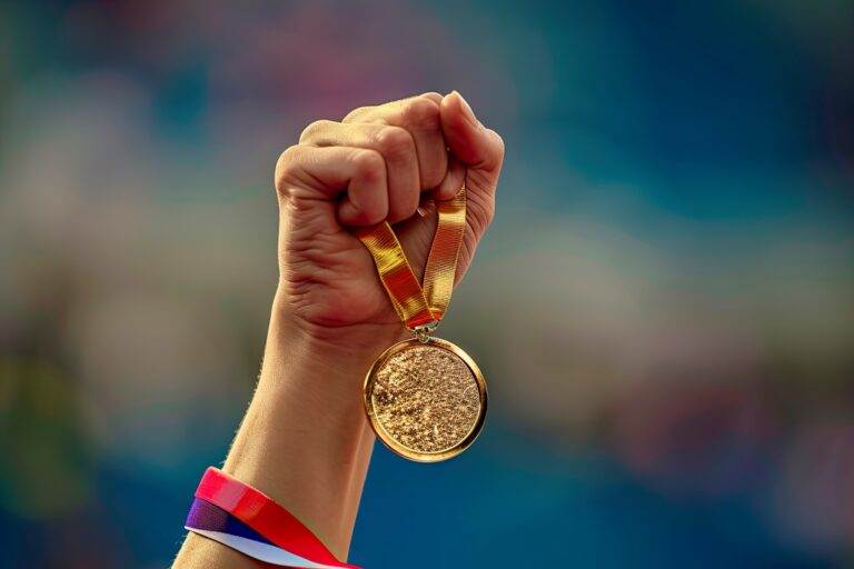 Une main serrée dans un poing brandit une médaille d'or attachée à un ruban, sur fond bleu flou, symbolisant la victoire et l'accomplissement aux Jeux Olympiques Paris 2024.